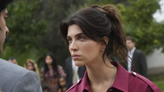 Close-up of a woman with dark brown hair in a ponytail, wearing a maroon jacket, outdoors, looking to the left with a neutral expression (blurred background).