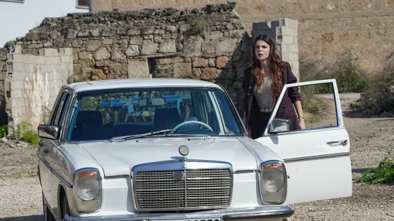 A woman with long wavy hair wearing a blazer stands next to an open driver’s door on a white vintage car in a rocky outdoor setting.